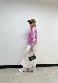 Ružová mikina na zips s bielymi pruhmi, biela čipkovaná sukňa, biele tenisky s ružovými akcentmi, zelená taška s logom a olivová čiapka so slnečnými okuliarmi.