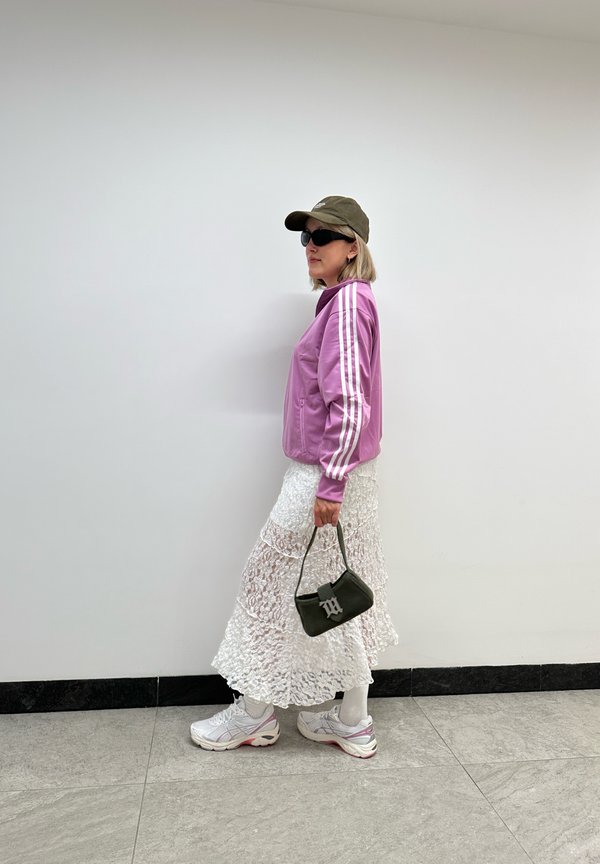 Rosa Zip-Jacke mit weißen Streifen, weißer Spitzenrock, weiße Sneakers mit rosa Akzenten, grüne Tasche mit Logo und olivgrüne Mütze mit Sonnenbrille.
