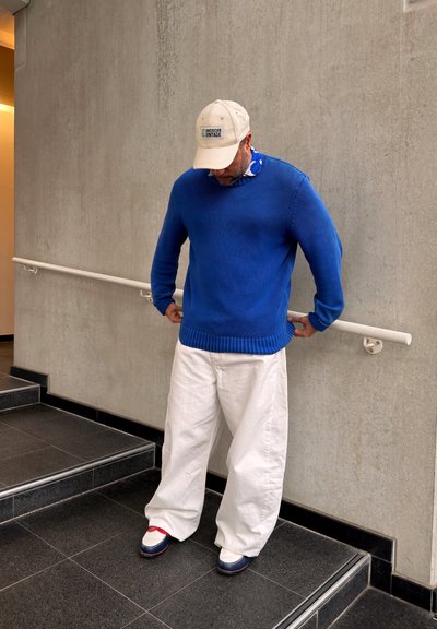 Hombre con suéter azul, pantalones anchos blancos, gorra beige y zapatillas, de pie junto a una barandilla junto a unas escaleras contra una pared gris.