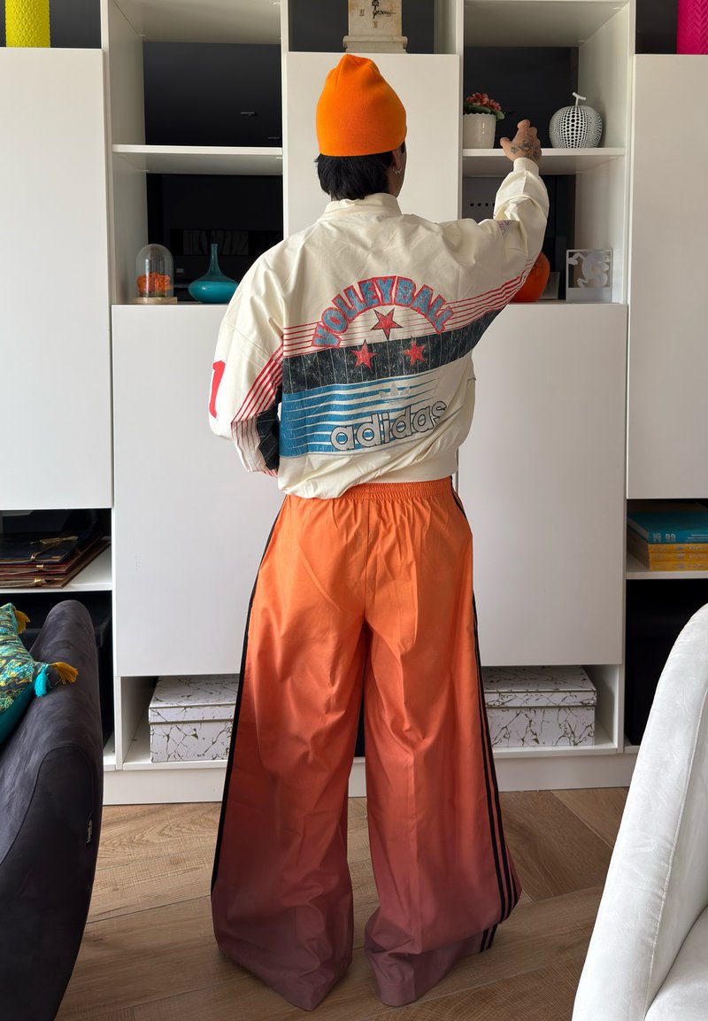 Personne portant un bonnet orange, une veste de volley-ball blanche Adidas et un pantalon large orange, atteignant un objet sur une étagère blanche dans un salon.