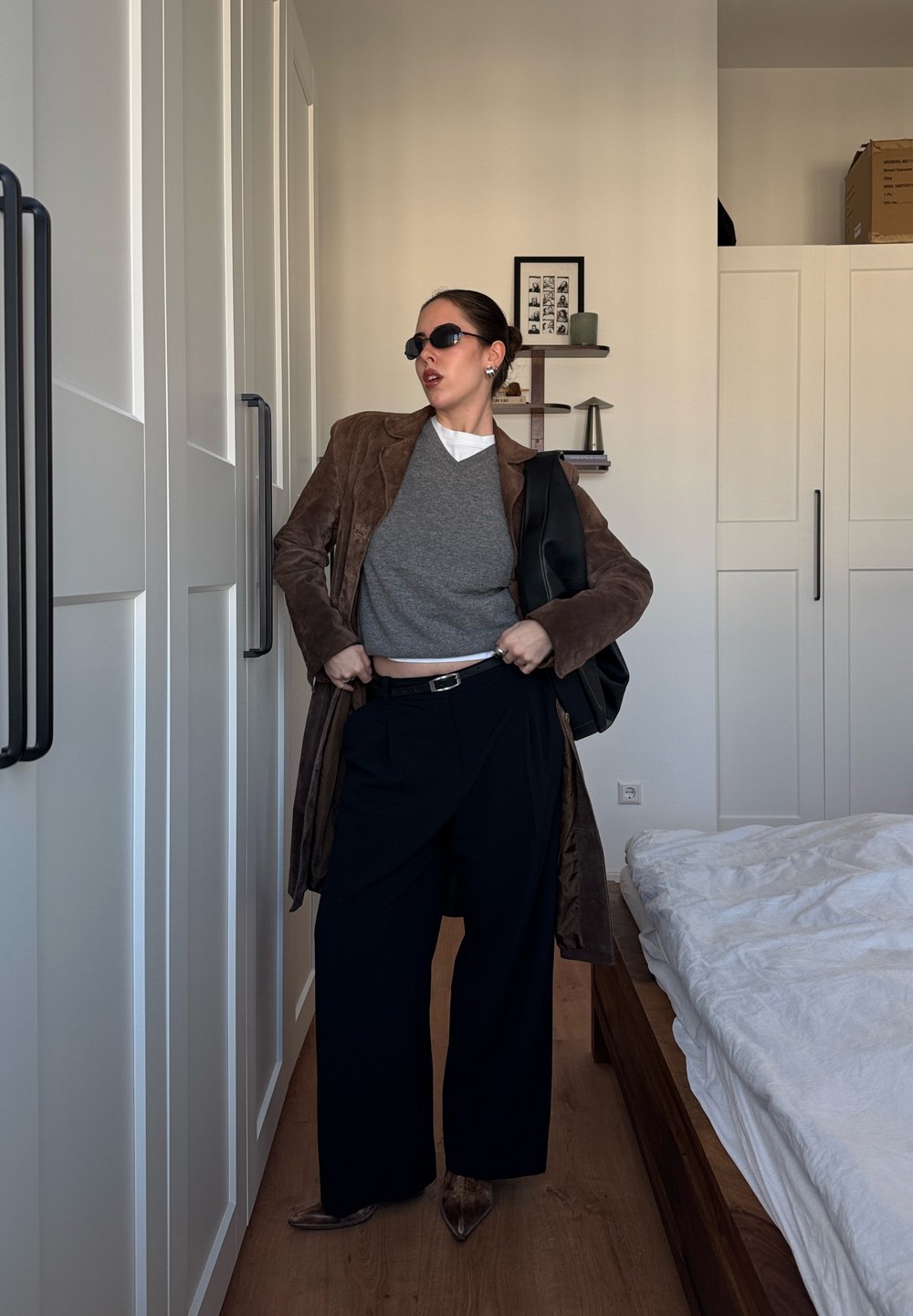Long manteau en daim marron porté sur un pull gris, pantalon large noir et chaussures marron à bout pointu. Sac noir et lunettes de soleil complètent le look.