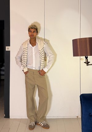 Persona joven con cabello rizado y rubio, de pie contra una pared blanca, vistiendo una camiseta blanca, un suéter a rayas colgado sobre los hombros, pantalones beige y zapatillas deportivas.