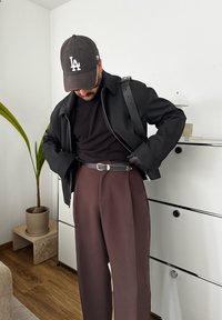 Μαύρο μπουφάν με φερμουάρ και λεία υφή, μαύρο T-shirt, καφέ παντελόνι ψηλής μέσης και μαύρο καπέλο με λογότυπο "LA", εντός του χώρου.