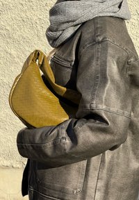 Mala de couro trançado em amarelo mostarda, com uma forma curva e textura suave. A modelo usa um cachecol cinza e uma jaqueta escura com fecho éclair.