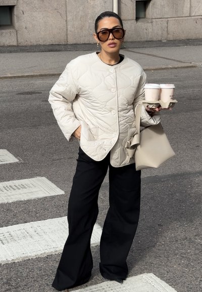 Chaqueta acolchada beige con botones redondos, pantalones de pierna ancha negros y un bolso beige. Sosteniendo dos tazas de café en un portavasos.