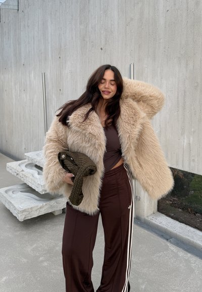 Abrigo de piel sintética en color beige claro, diseño oversize con capucha. Pantalones deportivos marrones con rayas blancas. Sosteniendo un bolso tejido de color verde oscuro.