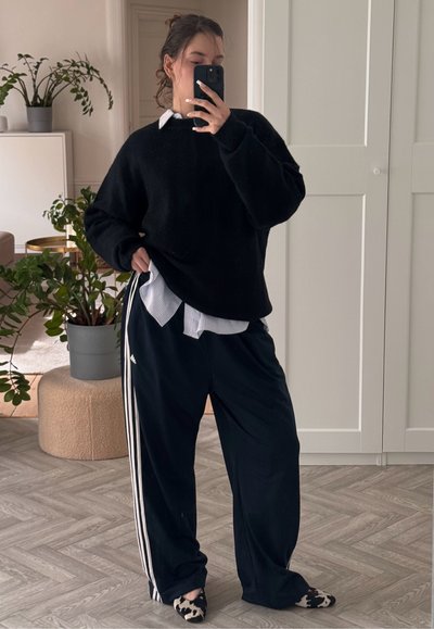 Suéter negro de punto de gran tamaño colocado sobre una camisa a rayas azul claro, combinado con pantalones negros holgados con rayas blancas y zapatos estampados.