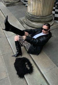 Mustat polveen ulottuvat korkokengät, joissa on kärjellinen varvas, yhdistettynä maastokuvioiseen hameeseen ja mustaan tekoturkiskaulaliinaan, harmaiden kivisten portaiden päällä ulkotilassa.