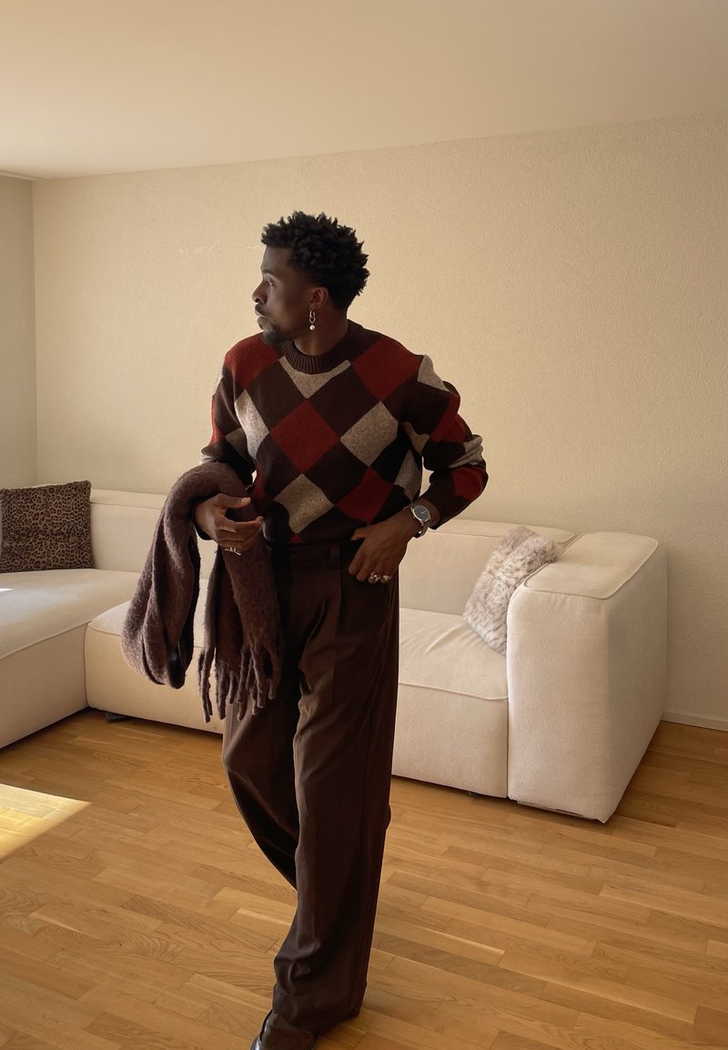 Homme portant un pull à motifs et un pantalon marron tenant une écharpe, debout dans un salon avec un canapé crème et un sol en bois.