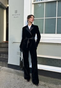 Femme en costume en velours noir avec revers et ceinture en satin, gants noirs, tenant un sac noir, debout devant un bâtiment blanc au 11 Penzance Place.