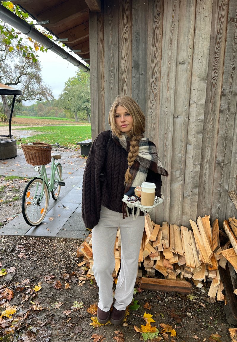 Une femme tenant un plateau avec du café et une pâtisserie se tient à côté d’un tas de bois et d’un vélo vert menthe, devant une cabane rustique en bois.