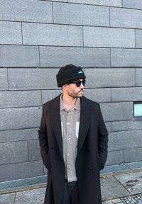 Cappotto di lana nero, camicia a righe con taschino bianco, berretto nero con logo e occhiali da sole; in piedi contro un muro grigio testurizzato.