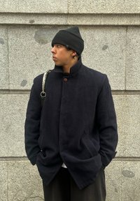 Μα Navy μάλλινο παλτό με ψηλό γιακά, δύο μπροστά τσέπες και ξύλινο κουμπί. Φορεμένο με μαύρο σκουφί και ένα μοτίβο στον λουρί.