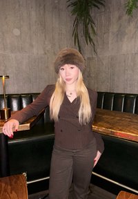 Brown pinstripe double-breasted jacket, matching brown trousers, and a fur hat; set against a dark green leather banquette and wooden table.