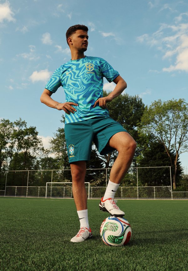 Jeune homme en tenue de football brésilienne bleue debout sur un terrain en herbe avec un pied sur un ballon de football coloré sous un ciel partiellement nuageux.