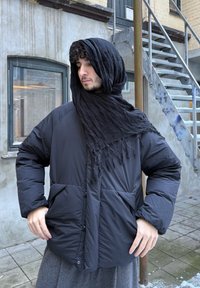 Doudoune noire avec de grandes poches, dotée d'une texture mate et de fermetures à boutons, associée à une écharpe noire à franges. Environnement extérieur.