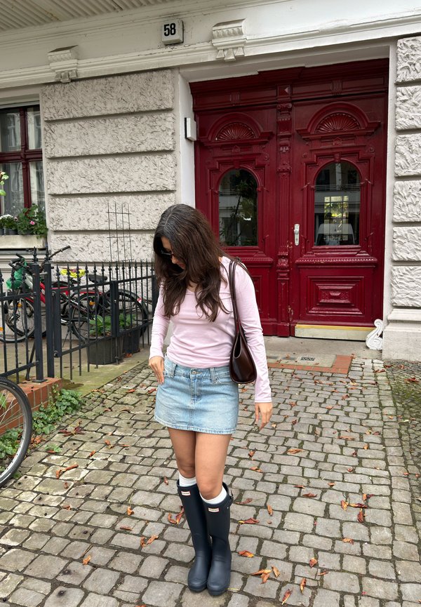 Mujer con top rosa, falda vaquera y botas de lluvia negras de pie en un camino empedrado frente a un edificio con grandes puertas dobles rojas.