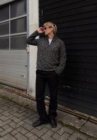 Siva jakna s leopardjim vzorcem in zadrgo, z dvignjenim ovratnikom, črne hlače in črne prešite čevlje. Stoji ob temnem lesene steni.