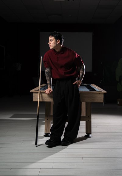 Hombre con tatuajes, vestido con una camisa roja y pantalones negros, se apoya en una mesa de billar mientras sostiene un taco en una habitación con poca luz.