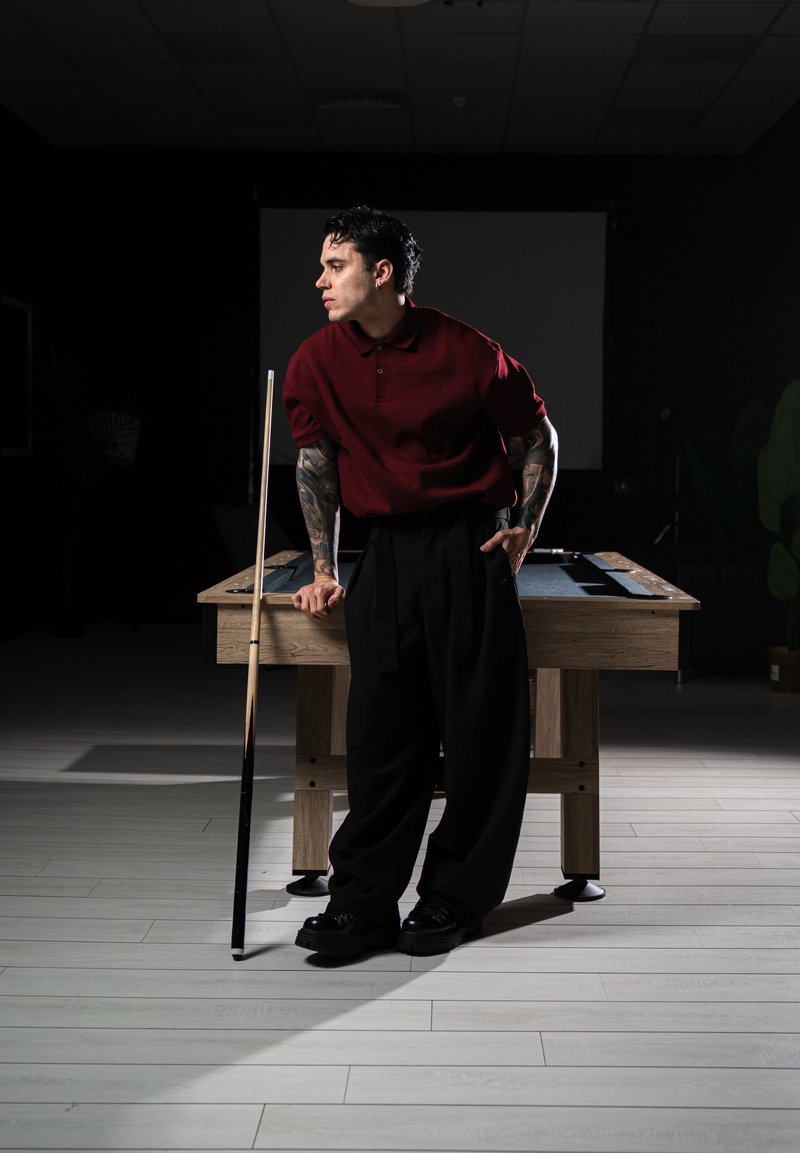 Homme tatoué portant une chemise rouge et un pantalon noir, appuyé sur une table de billard en tenant une queue de billard dans une pièce faiblement éclairée.