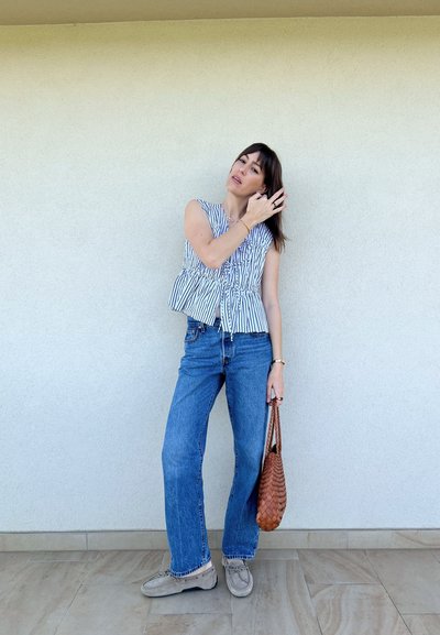 Top peplum sin mangas a rayas en blanco y azul, combinado con jeans azul claro. Los accesorios incluyen un bolso de mimbre marrón y calzado beige.