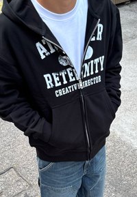 Fekete zipzáros kapucnis pulóver fehér grafikával, rajta a "RETERNITY" és a "CREATIVE DIRECTOR" feliratokkal. Fehér póló fölött viselve, az elülső zsebek láthatóak.