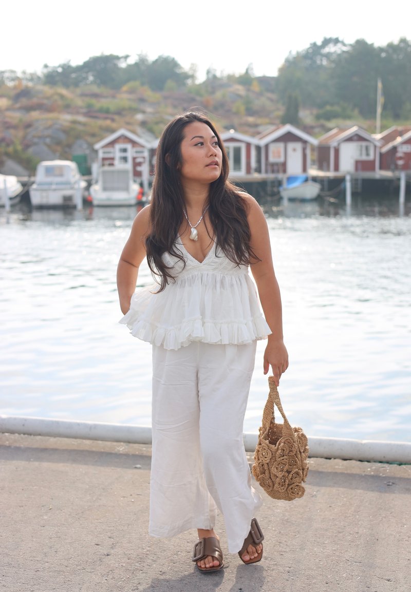 Top bianco arricciato abbinato a pantaloni bianchi a gamba larga, sandali marroni a scivolo e una borsa all'uncinetto beige con dettagli floreali. Acqua e barche sullo sfondo.