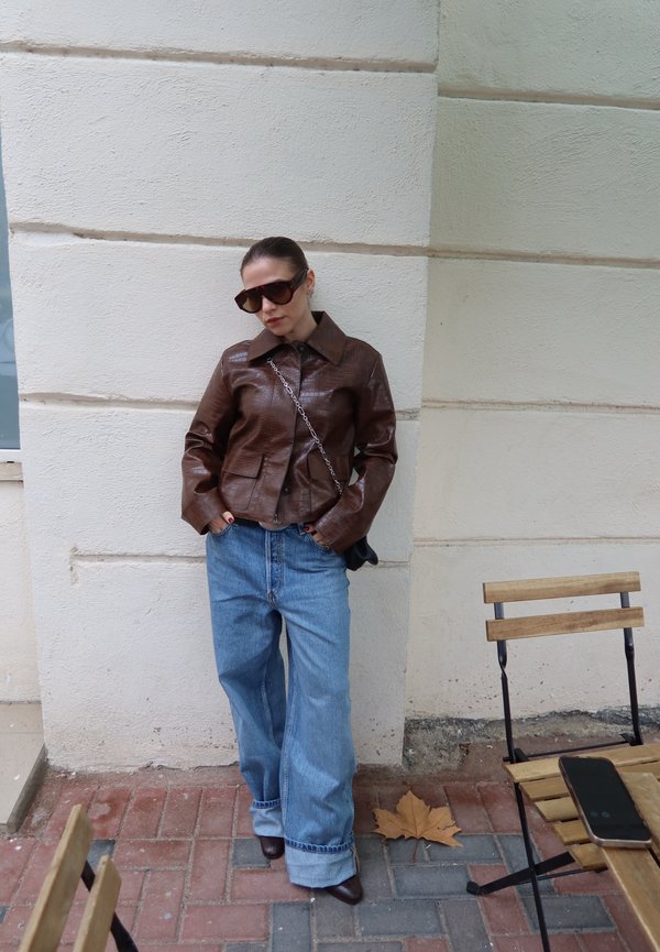 Une femme portant des lunettes de soleil, une veste en cuir marron et un jean bleu ample pose contre un mur en pierre claire, à côté d’une chaise en bois.