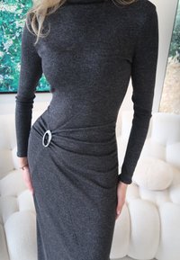 Γυναίκα που φοράει εφαρμοστό μακρυμάνικο σκούρο γκρι φόρεμα με ασημέν ια αγκράφα στη μέση, στέκεται μέσα σε εσωτερικό χώρο κοντά σε λευκά έπιπλα.