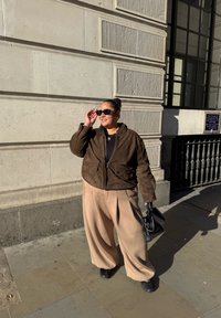 Casaco de camurça castanho, blusa preta, calças largas beje, sapatos pretos, segurando uma pequena bolsa preta. Os acessórios incluem óculos de sol e um colar prateado.