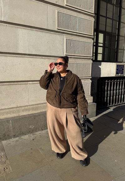 Chaqueta de ante marrón, blusa negra, pantalones de pierna ancha beige, zapatos negros, sosteniendo un pequeño bolso negro. Los accesorios incluyen gafas de sol y un collar de plata.