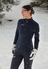 Veste zippée marine avec accents orange, associée à un pantalon assorti et des gants couleur crème. Fond enneigé qui met en valeur le thème des sports d'hiver.