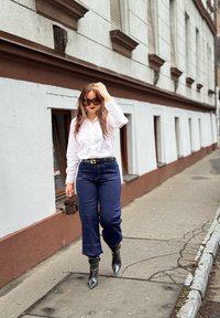 Femme marchant sur le trottoir en ville, portant une chemise blanche, un jean bleu foncé, des bottes noires, des lunettes de soleil, et portant un petit sac à main marron.