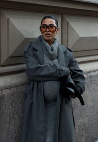 Femeie îmbrăcată într-o haină gri supradimensionată și o rochie pulover, purtând ochelari de soare mari cu nuanță, ținând o geantă neagră, stând lângă un perete din piatră texturată.