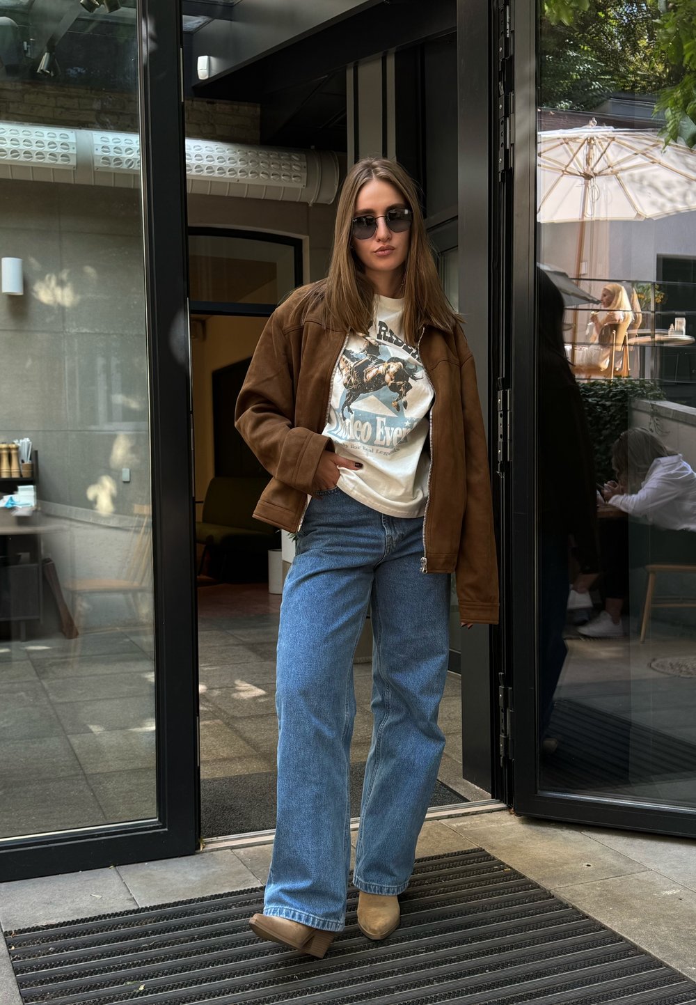 Jaqueta de camurça castanha sobre uma t-shirt gráfica, calças jeans largas de azul claro e sapatos de plataforma bege; de pé junto a uma entrada de vidro.