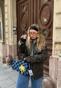 Noor naine, pikkade laineliste juustega, must peapael, prillid, nahktagi, hoiab väljas sinist ja pruunikat ruudulist kotti kollase palmimotiiviga.