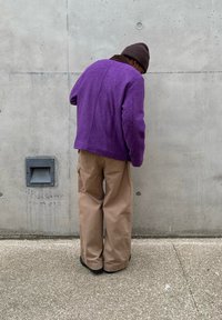 Personne portant une veste violette, un pantalon large couleur beige et un bonnet marron, debout et se penchant en avant contre un mur en béton uni.