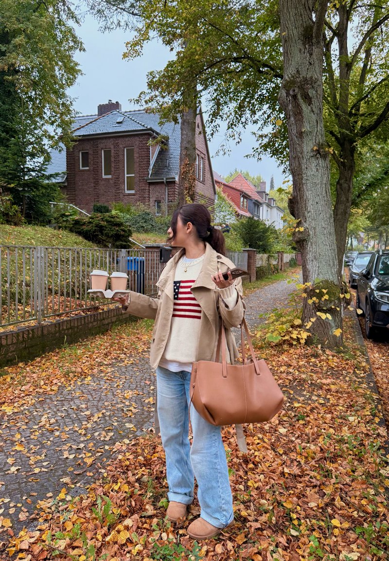 Tan jacket over a cream sweater with red and navy stripes, blue jeans, brown handbag, holding two coffee cups, surrounded by autumn leaves.