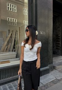 Top bianco senza spalle con dettaglio arriccicato, pantaloni neri a vita alta e occhiali da sole, completato da un accessorio per capelli a forma di rosa.