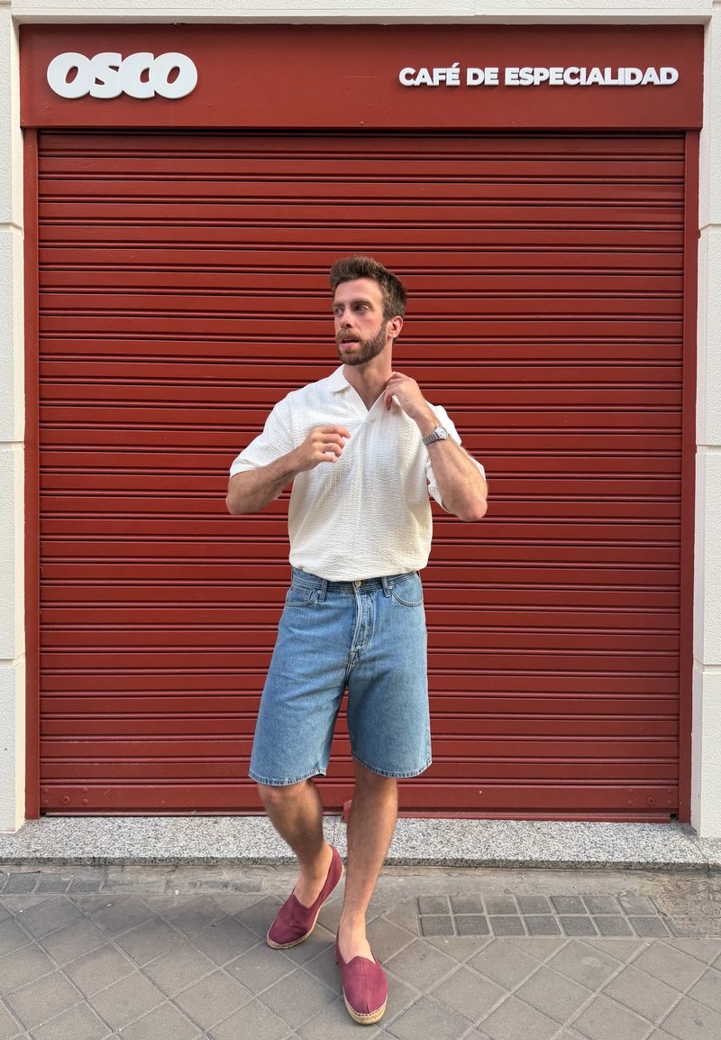 Homme en chemise blanche, short en jean et chaussures rouges, debout devant un volet métallique rouge fermé avec une enseigne de café au-dessus.