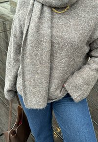 Un pull en laine marron avec une finition texturée, associé à un jean bleu. Un accent de chaîne en or est visible près de l'encolure. Un sac marron est tenu.