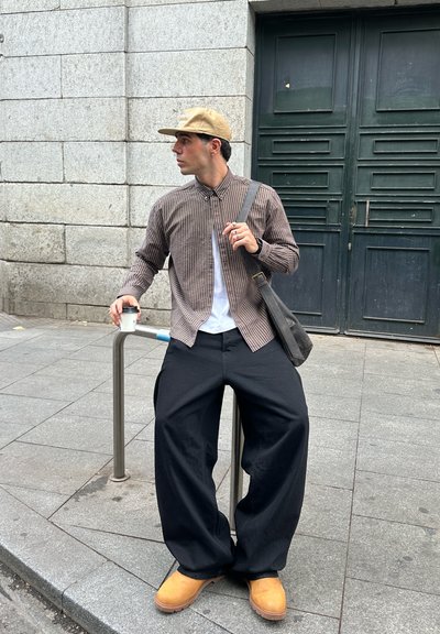 Camisa de cuadros marrón y blanca, camiseta blanca, pantalones negros de pierna ancha, zuecos color canela, gorra caqui, sosteniendo una taza de café, de pie junto a una barandilla.