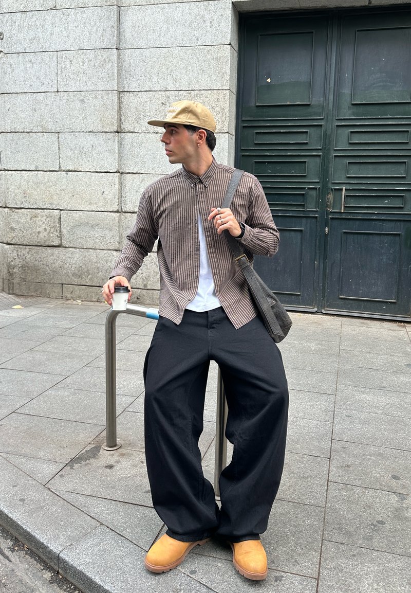 Chemise à carreaux marron et blanche, T-shirt blanc, pantalon large noir, sabots marron clair, casquette kaki, tenant une tasse de café, debout près d'une rambarde.