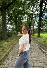 Sweater de malha em creme de mangas curtas combinado com jeans de perna larga cinzentos. Caminho de pedra ladeado por árvores e vegetação ao fundo.