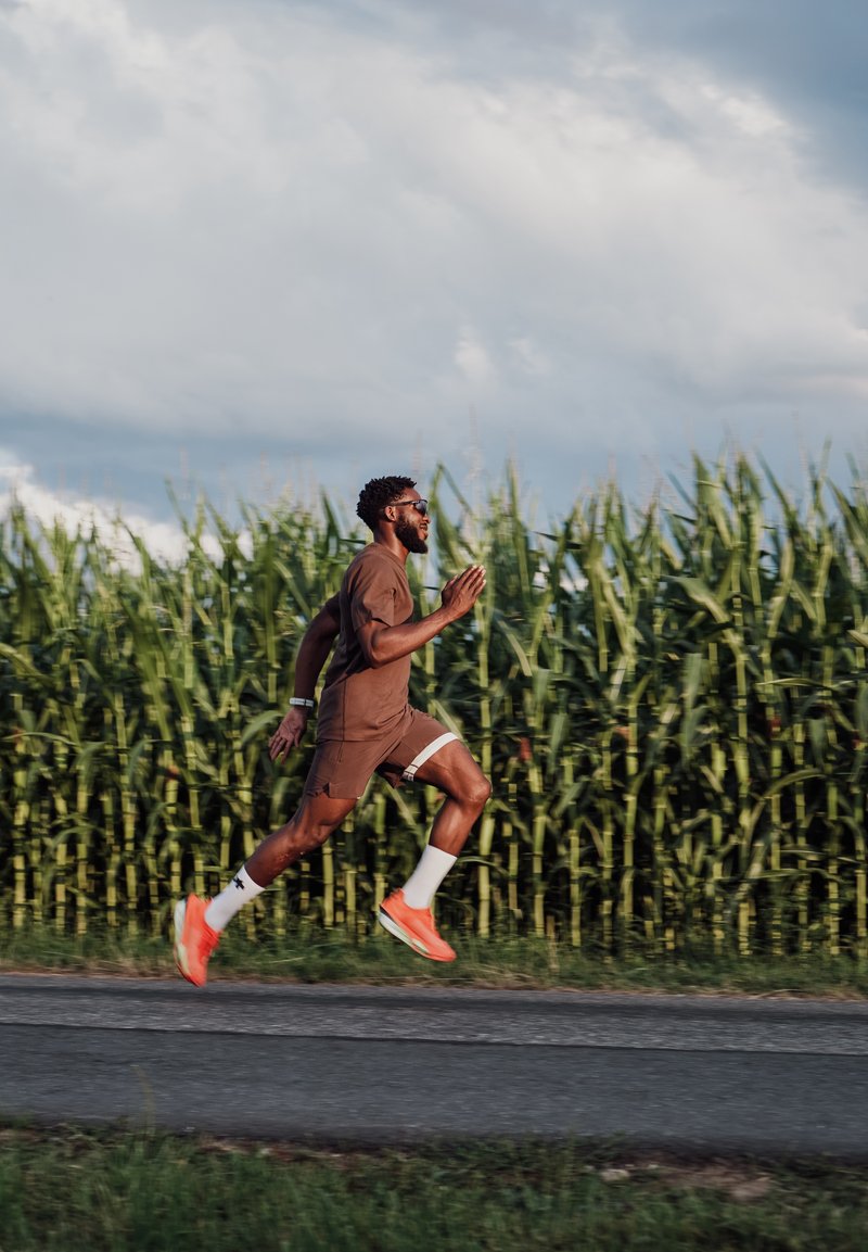 Homme portant des vêtements de sport marron et des chaussures orange courant sur une route à côté d'un grand champ de maïs vert sous un ciel nuageux.