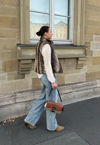 Colete de peluche, camisa de manga longa branca, calças largas de ganga azul claro, botas até ao tornozelo em verde-oliva e uma mala castanha com detalhes pretos, a caminhar.