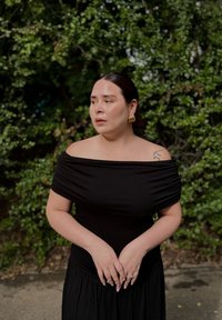 Robe noire à épaules nues avec texture froncée ; le modèle porte des boucles d'oreilles en or et un petit tatouage sur l'épaule, le tout au milieu d'une verdure.