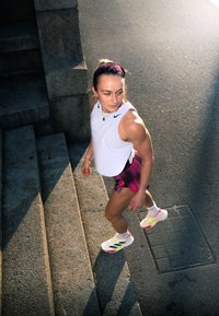 Atlet v bílém bezrukávovém tričku a vzorovaných růžových šortkách, obutý v bílých a barevných běžeckých botách, schází po kamenných schodech.