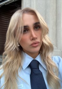 Lyshåret person med bølget hår iført en blå skjorte med krave og et mørkt slips med mønster, stående udendørs nær en bygning med stenvægge.