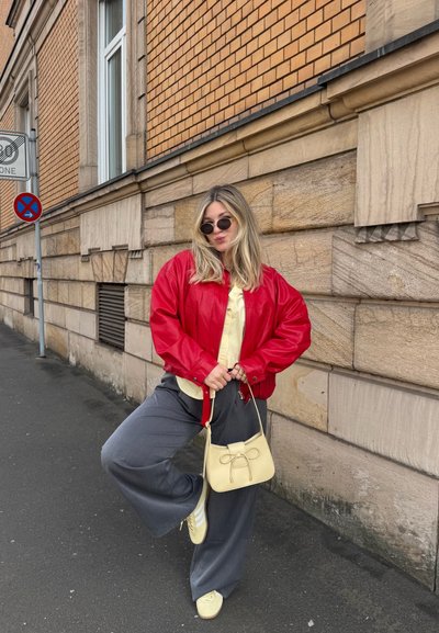 Joven mujer con gafas de sol que lleva una chaqueta roja, camisa amarilla y pantalones grises, está de pie en la calle frente a una pared de ladrillos sosteniendo un bolso beige.
